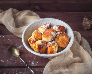 Homemade cheesecakes in a deep white plate sprinkled with powdered sugar. A beige napkin and a teaspoon with a twisted handle complete the composition.