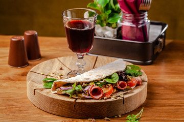 Concept Italian cuisine. Piadina with ham, tomatoes, mix lettuce, pistachios, cucumbers on wooden board. Glass of red wine on the table. Beautiful serving dishes in the restaurant.
