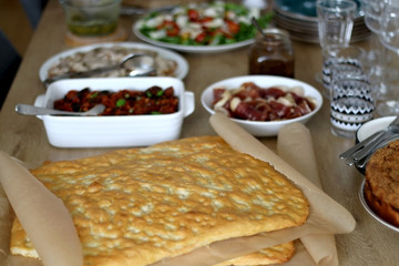Lavish homemade lunch served on table: focaccia bread, prosciutto, porchetta, mozzarella cheese, pesto, caponata, caramelized onion, with arugula and cherry tomato salad. Selective focus.