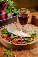 Concept Italian cuisine. Piadina with ham, tomatoes, mix lettuce, pistachios, cucumbers on wooden board. Glass of red wine on the table. Beautiful serving dishes in the restaurant.