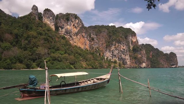 Traditional long-tailed Asian boat bobs on calm waves of Andaman Bay near Railay Beach, Krabi. Large rocky cliff with green vegetation and blue sky in background. 50 frames per second full hd footage.