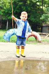 A three year old boy in yellow rubber boots and a denim blue shirt stands with a stick near a puddle
