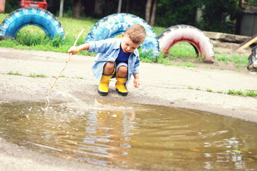 A three-year-old boy in yellow rubber boots and a denim blue shirt plays with a stick in a puddle