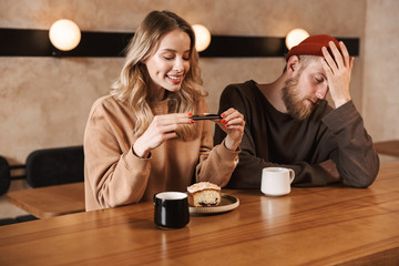 Happy young woman take photo of breakfast nearher displeased man in cafe drinking coffee.