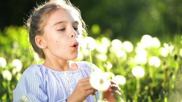 Beautiful little girl at sunny summer day blowing dandelion on green meadow. Summer fun concept. Slow motion.