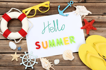 Word Hello Summer with seashells, lifebuoy and flip flops on brown wooden table