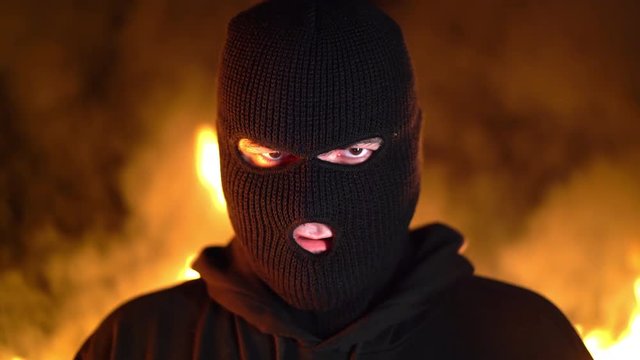 Portrait of young man in black mask during street fights against burning fire background. Concept of political strikes and football hooligans - ultras