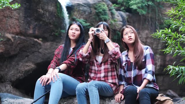 A group of friends, women, teenagers, tourists from Asia, with three people sit and take pictures of natural scenery on the waterfall in the forest. Adventure travel holidays. Slow motion