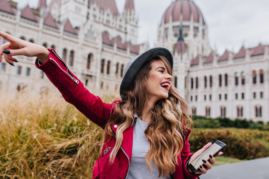 Carefree caucasian girl in trendy red jacket fooling around during european trip. Excited young lady in hat exploring city and dancing with amazing building on background.