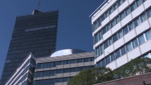 The Kista Science Tower and other buildings with walls of reflective glass and sleek, modern design. Trees behind a brick wall blow in a gentle summer breeze. Bright blue sky and tall office building.