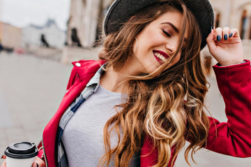 Close-up portrait of fascinating white woman with golden wavy hair posing with eyes closed on the street. Romantic relaxed girl in hat holding cup of latte. - obrazy, fototapety, plakaty