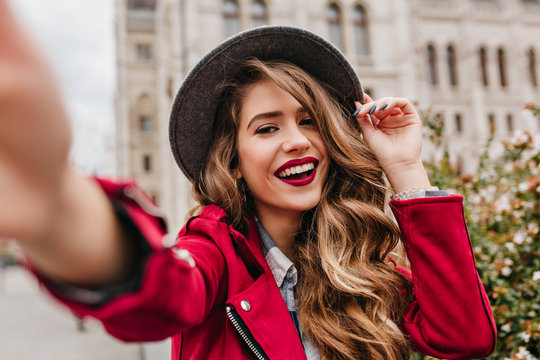 Portrait of wonderful white female model with bright makeup expressing energy in good day in Europe. Lovely curly woman in stylish hat making selfie while walking past old building.