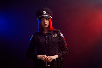 Serious female police officer is posing for the camera against a black background with red and blue backlighting.
