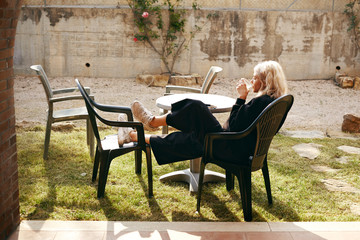 Fototapeta na wymiar Outdoor portrait of fashionable pretty blonde girl in her twenties chilling outside house enjoying morning coffee and gentle sunshine sitting at round table, holding mug, keeping feet on chair