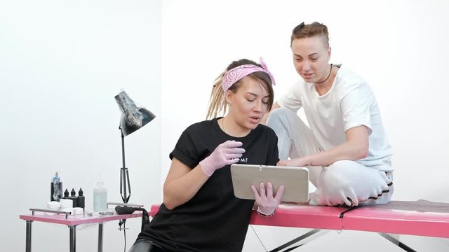 Tattoo master woman discusses sketch with client, shows pictures on tablet in studio. Pink background.