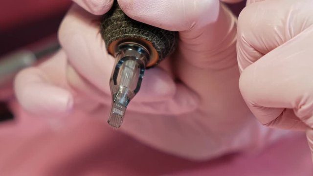 The tattoo master inserts a needle into the tattoo machine. Pink safety gloves on a red background. Female master artist.