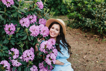 Beautiful young woman with long brunette hair in straw hat posing near pink flowers in a garden