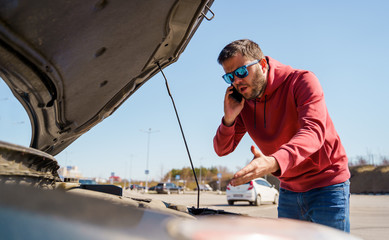 Photo of upset man with phone in next to open hood of broken car in daytime