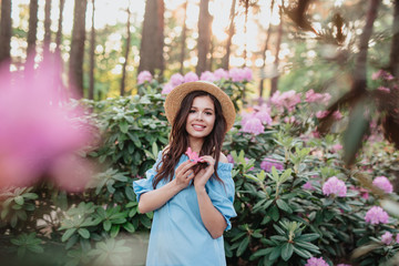 Beautiful girl in blue vintage dress and straw hat standing near pink flowers.