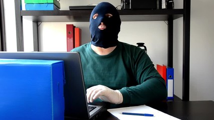 Thief inserts USB flash memory stick into a laptop in robbed office. He wears a black balaclava and medical latex gloves