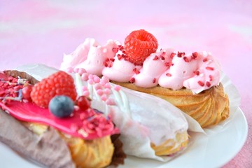 Tasty French eclairs cake with creative pink decor and fresh berries on pink textured table. Selective focus. Delicious dessert profiteroles with pink and red icing and sugar decor elements. 