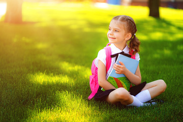 Little school girl with pink backpack sitting on grass after lessons looking side away, read book or study lessons, thinking ideas, education and learning concept.