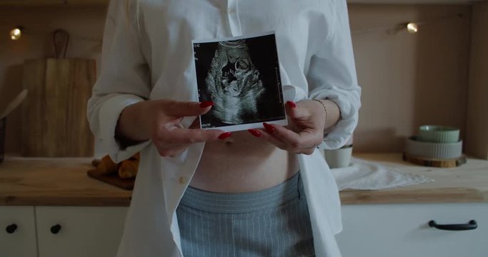 Caucasian pregnant women with big belly standing in kitchen and holding in front of her sonogram image. 60 fps