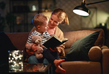 Family before going to bed mother reads to her child son book near a lamp in the evening - obrazy, fototapety, plakaty