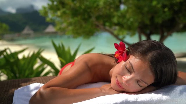 Spa Luxury Woman relaxing getting massage at hotel resort. Beauty girl lying down sleeping on towel, hibicus flowers outside. Serene ethnic woman relaxing. Mixed race Asian / Caucasian model outdoor