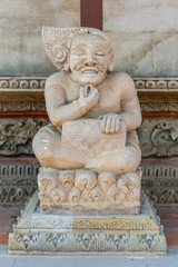 Ubud, Bali, Indonesia - February 26, 2019: Batuan Temple. White stone thinking man statue in front of shrine wall with brown beam, and decorated stone base.