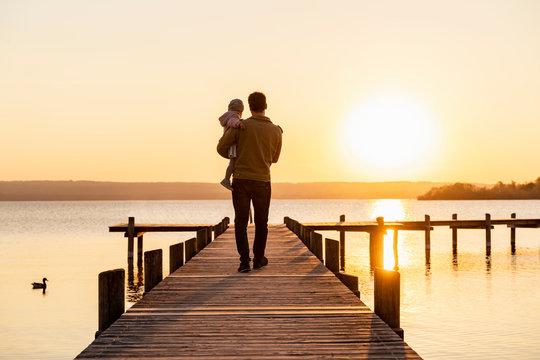 Germany, Bavaria, Herrsching, father carrying daughter on jetty at sunset
