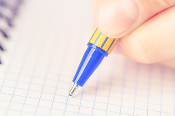 A man or teenager writes a blue-yellow pen in a checkered notebook.