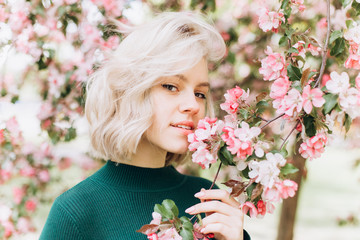 Fototapeta premium Close up portrait of young pretty caucasian curly blondie model posing outdoors in pink blossom garden. Beauty, nature concept