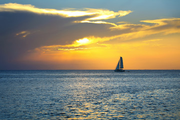 Colorful seascape image with shiny sea and sailboat over cloudy sky and sun during sunset in Cozumel, Mexico