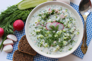 Okroshka is a traditional Russian summer cold soup in a ceramic bowl. Ingredients potatoes, radishes, cucumbers, dill, eggs, kefir yogurt, sausage. Summer yogurt cold soup on a wooden table. Selective