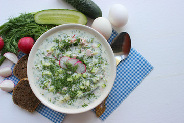 Okroshka is a traditional Russian summer cold soup in a ceramic bowl. Ingredients potatoes, radishes, cucumbers, dill, eggs, kefir yogurt, sausage. Summer yogurt cold soup on a wooden table. Selective