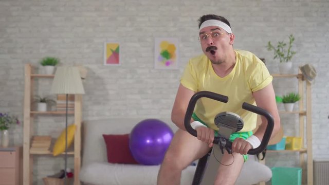 Young funny man athlete from the 80's with a mustache falls off a exercise bike