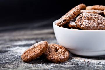 Fresh baked chocolate chip and oat fresh cookies with sugar powder heap in white bowl on black background.