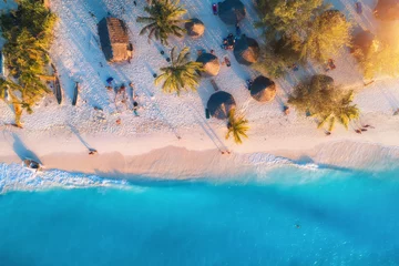Fotobehang Luchtfoto van parasols, palmen op het zandstrand van de Indische Oceaan bij zonsondergang. Zomer reizen in Zanzibar, Afrika. Tropisch landschap met palmbomen, parasols, mensen, zand, blauw water, golven. Bovenaanzicht © den-belitsky