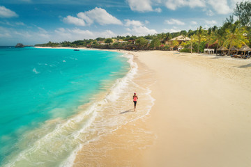 Aerial view of the running young woman on the white sandy beach near sea with waves at sunny day....