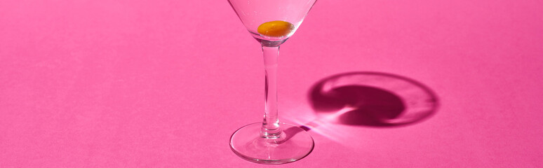 panoramic shot of transparent glass with cocktail and olive on pink background