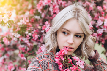 Fototapeta premium Close up portrait of young pretty caucasian curly blondie model posing outdoors in pink blossom garden. Beauty, nature concept