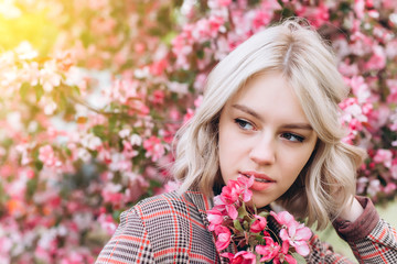 Fototapeta premium Close up portrait of young pretty caucasian curly blondie model posing outdoors in pink blossom garden. Beauty, nature concept