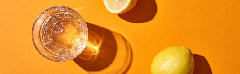 top view of glass with water near yellow lemons on orange background, panoramic shot