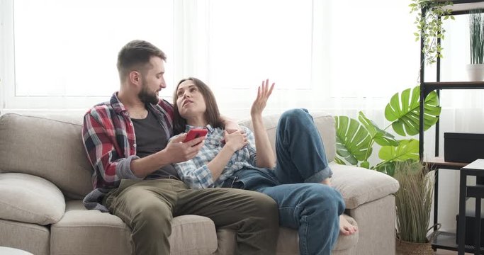 Relaxed couple using mobile phone and talking with each other while lying on sofa at home