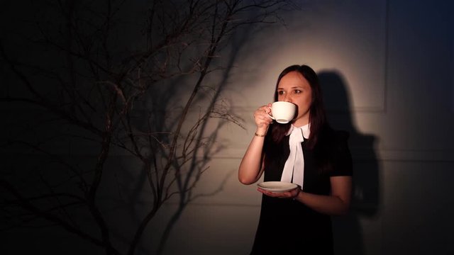A woman in an elegant dress drinking coffee in a dark room. Sensual female with hair style holding a cup of coffee in elegant blak dress over dark fashion background