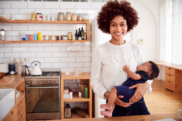 Portrait Of Smiling Mother Holding Sleeping Baby Son In Kitchen