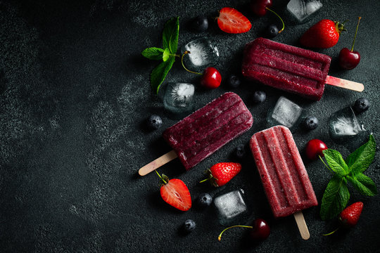 Bright popsicle made of cherries, strawberries and blueberries on a dark concrete background. Sweet summer treat. Top view with copy space. Flat lay
