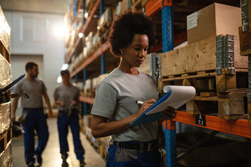 Black female warehouse worker writing delivery schedule list.