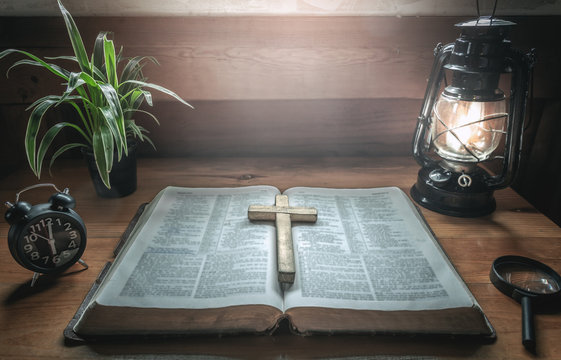 Close up wooden christian cross putting on old holy Bible with light of oil lamp at worship room. christian concept.